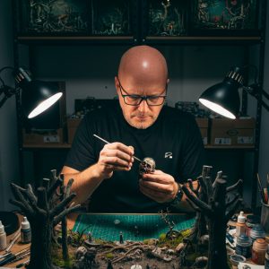 Víctor M., artesano de 27dioramas, pintando a mano una miniatura de calavera en su taller rodeado de dioramas artesanale